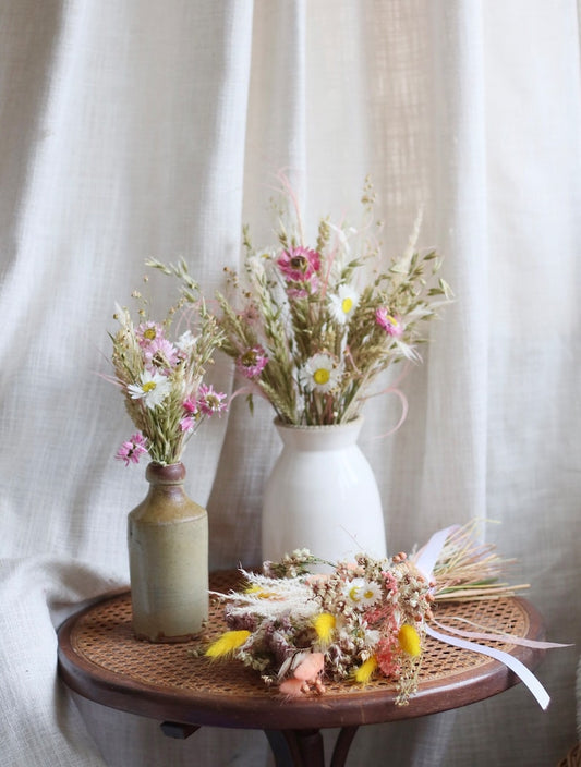 Care guide how to care for dried flowers. 3 flower bouquets in neutral toned vintage vases on a cane wooden table displayed for a photograph